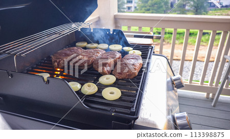 The outdoor two-burner gas grill is put to good use, sizzling with the sound and aroma of ribeye steaks and onion rings being perfectly cooked. 113398875