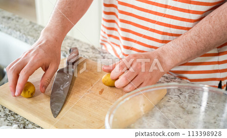 In the contemporary ambiance of a modern kitchen, a young man engages in dinner preparations. His current activity entails meticulously slicing small rainbow potatoes in half on a wooden cutting board 113398928