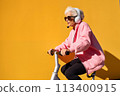 Smiling happy cool gray haired old senior woman on scooter or bicycle wearing headphones with a microphone. The Hands free concept 113400915