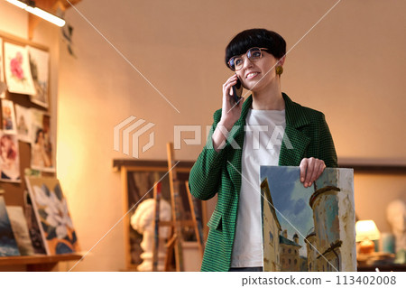 Waist up portrait of smiling female artist speaking on phone and holding painting in workshop, copy space 113402008