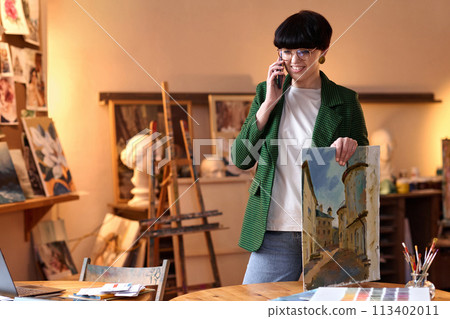 Waist up portrait of female art expert speaking on phone and holding painting in gallery shop 113402011