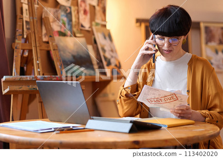 Portrait of woman holding Past due bill and speaking on phone in art studio, copy space Portrait of woman holding Past due bill and speaking on phone in art studio, copy space 113402020