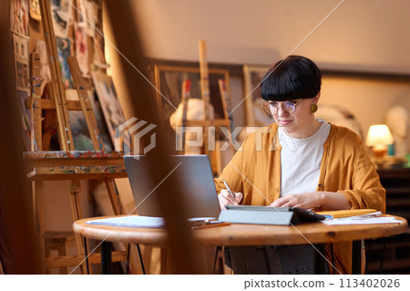 Portrait of creative adult woman using laptop in art studio and studying in online lesson Portrait of creative adult woman using laptop in art studio and studying in online lesson 113402026