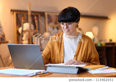 Portrait of smiling female artist using laptop in studio giving online art class and speaking via video chat Portrait of smiling female artist using laptop in studio giving online art class and speaking via video chat 113402031