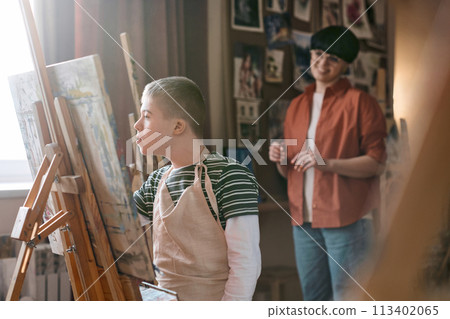 Soft side view portrait of young boy with Down syndrome painting on easel in art therapy class with mentor watching copy space Soft side view portrait of young boy with Down syndrome painting on easel in art therapy class with mentor watching copy space 113402065