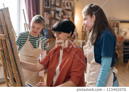 Side view portrait of smiling young woman painting on easel in art class with children watching 113402078