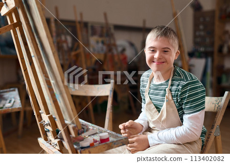 Portrait of young boy with Down syndrome smiling at camera sitting by easel in art class copy space 113402082