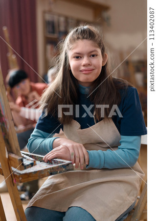 Vertical portrait of smiling young girl with disability sitting by easel in art class and looking at camera 113402087