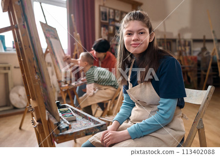 Portrait of teen girl with disability smiling at camera sitting by easel in art class copy space 113402088