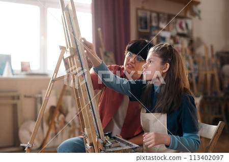 Side view portrait of young girl with disability painting on easel in art class with female teacher assisting copy space Side view portrait of young girl with disability painting on easel in art class with female teacher assisting copy space 113402097