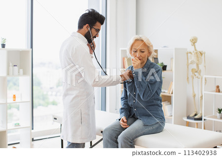 Bearded man in white lab coat checking lungs and heart via stethoscope. Bearded man in white lab coat checking lungs and heart via stethoscope. 113402938