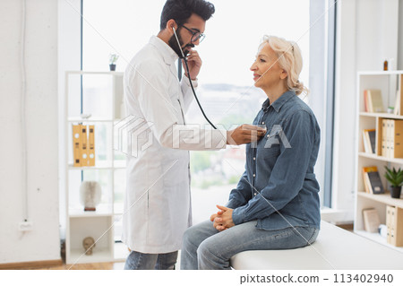 Adult man using stethoscope while elderly woman sitting on exam couch. 113402940