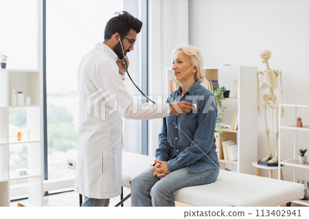 Adult man using stethoscope while elderly woman sitting on exam couch. 113402941