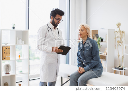 Bearded male doctor and elderly female patient sitting at exam couch. Bearded male doctor and elderly female patient sitting at exam couch. 113402974