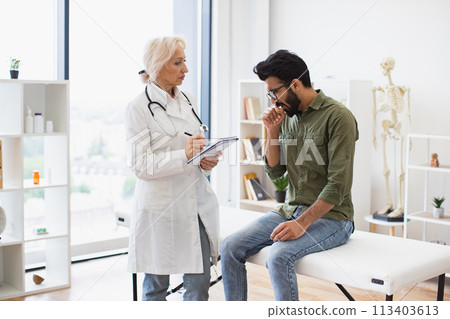 Caucasian senior female doctor and young male patient sitting at exam couch. 113403613