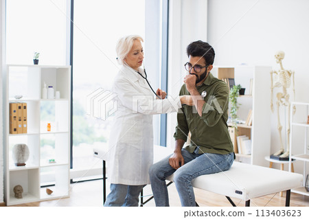 Elderly lady using stethoscope while young man sitting on exam couch. Elderly lady using stethoscope while young man sitting on exam couch. 113403623