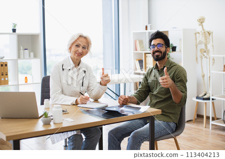 Portrait of happy female senior doctor and young male patient sitting at table. 113404213
