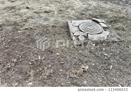A rusty iron hatch with a pattern and a large gap at the top is mounted in a broken cracked asphalt structure well next to the road. Close-up 113406015