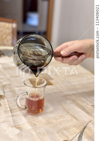 A woman's hand pours delicious dark tea from a glass teapot into a glass vessel standing on the table. Close-up A woman's hand pours delicious dark tea from a glass teapot into a glass vessel standing on the table. Close-up 113406021