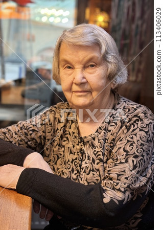 Portrait of positive mature old woman with gray hair aged 80s dressed in a brown jacket sitting at the table at the restaurant 113406029