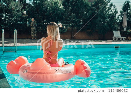 A woA woman is sitting on a pink inflatable flotation device in a pool. Concept of fun and relaxation, as the woman is enjoying her time in the water. 113406082