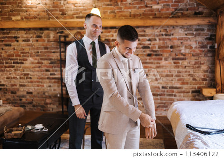 Handsome groom wears suit and groomsman helps him. Wedding morning. Background of brick wall Handsome groom wears suit and groomsman helps him. Wedding morning. Background of brick wall 113406122