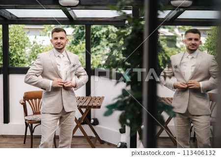 Young groom fastens the button on his jacket. Reflection of a man in a window Young groom fastens the button on his jacket. Reflection of a man in a window 113406132