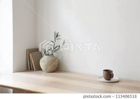 Breakfast, scandi indoor interior still life. Minimal home design concept. Beige ceramic vase with olive tree branches. Cup of coffee, tea on wooden table, desk with old books, empty beige wall mockup 113406518