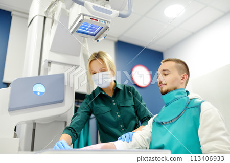 Female radiologist going to made x-ray shot of young man hand in x-ray room in modern clinic. Patient wearing in protect lead apron 113406933