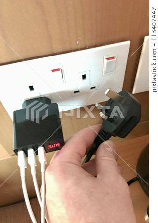 The man's hand holds a triple mains plug to connect a phone charger in the hotel. Nearby, the power supply is plugged into a double outlet The man's hand holds a triple mains plug to connect a phone charger in the hotel. Nearby, the power supply is plugged into a double outlet 113407447