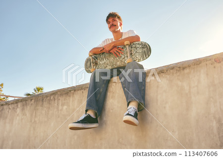 Smiling young male skateboarder holding skateboard sitting on ramp in skate park 113407606