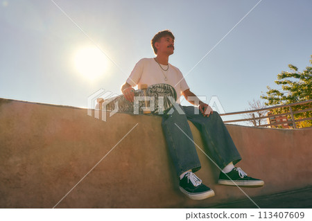 Smiling young male skateboarder holding skateboard sitting on ramp in skate park 113407609