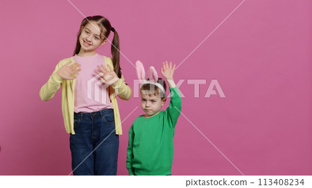 Joyful little kids waving in front of camera during easter holiday, smiling and wearing bunny ears. Brother and sister toddlers greeting someone in studio, adorable happy children. Camera A. 113408234