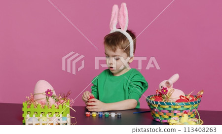 Adorable little youngster creates handcrafted ornaments for Easter Sunday and painting eggs with stamps and brushes. Pleased young boy using art materials to color and decorate. Camera A. 113408263