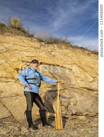senior male paddler wearing life jacket with a wooden canoe paddle on a rocky lake shore 113408400