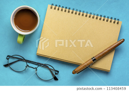 blank spiral notebook with reading glasses, pen and coffee on blue paper background 113408459