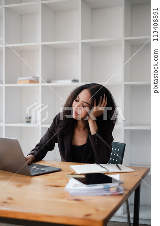 Businesswoman appears stressed as she leans on her hand, looking at her laptop in a modern office environment. 113408891