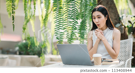Surprised and excited young woman at a laptop in a green, leafy outdoor cafe, with coffee and notebook on her table. Surprised and excited young woman at a laptop in a green, leafy outdoor cafe, with coffee and notebook on her table. 113408944
