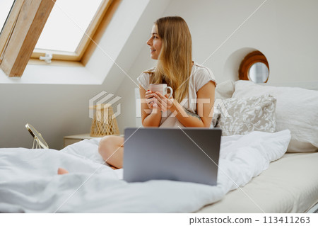 Woman sits on bed with laptop and coffee, relaxes with cozy interior design 113411263