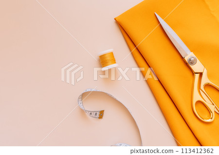 Sewing scissors lie on an orange fabric next to threads and a measuring tape Sewing scissors lie on an orange fabric next to threads and a measuring tape 113412362