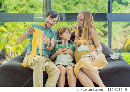 Heartwarming family moment as expectant mom, dad, and son joyfully browse through newborn baby's clothes, eagerly anticipating the arrival of a new family member 113415349