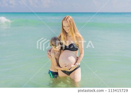 A picturesque moment: a pregnant woman and her son enjoying the turquoise sea, a heartwarming family bond by the tranquil shore A picturesque moment: a pregnant woman and her son enjoying the turquoise sea, a heartwarming family bond by the tranquil shore 113415729