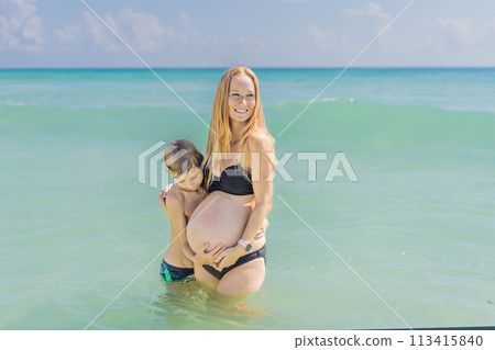 A picturesque moment: a pregnant woman and her son enjoying the turquoise sea, a heartwarming family bond by the tranquil shore A picturesque moment: a pregnant woman and her son enjoying the turquoise sea, a heartwarming family bond by the tranquil shore 113415840