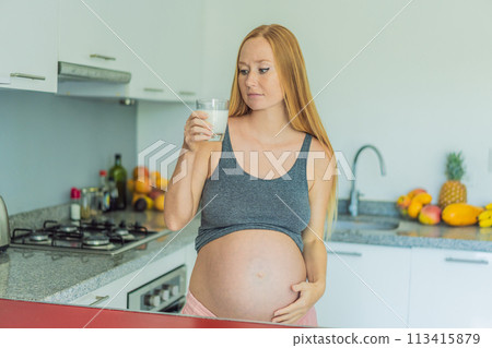 Weighing the pros and cons of milk during pregnancy, a thoughtful pregnant woman stands in the kitchen with a glass, contemplating the decision to include or avoid milk for her and her baby's well 113415879