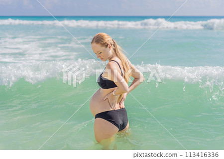 In the sea, a pregnant woman experiences a moment of discomfort as she stands, gently tending to her back, highlighting the challenges of back pain during pregnancy 113416336