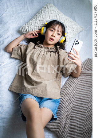 一個戴著耳機並在床上使用智慧型手機的女孩 一個戴著耳機並在床上使用智慧型手機的女孩 113417906