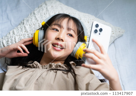 一個戴著耳機並在床上使用智慧型手機的女孩 一個戴著耳機並在床上使用智慧型手機的女孩 113417907