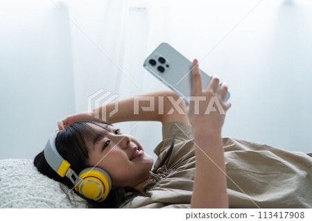 一個戴著耳機並在床上使用智慧型手機的女孩 113417908