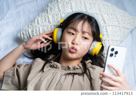 一個戴著耳機並在床上使用智慧型手機的女孩 113417913