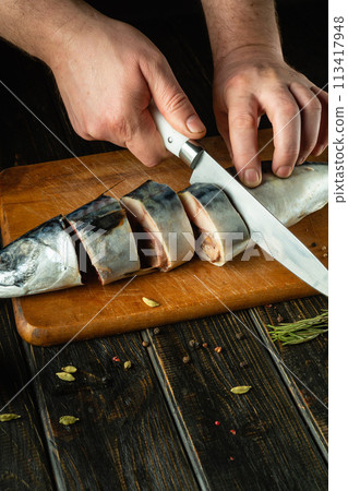 Slicing raw fish for salting with spices with a knife in the hands of a cook. The concept of preparing herring with fish on the kitchen table 113417948